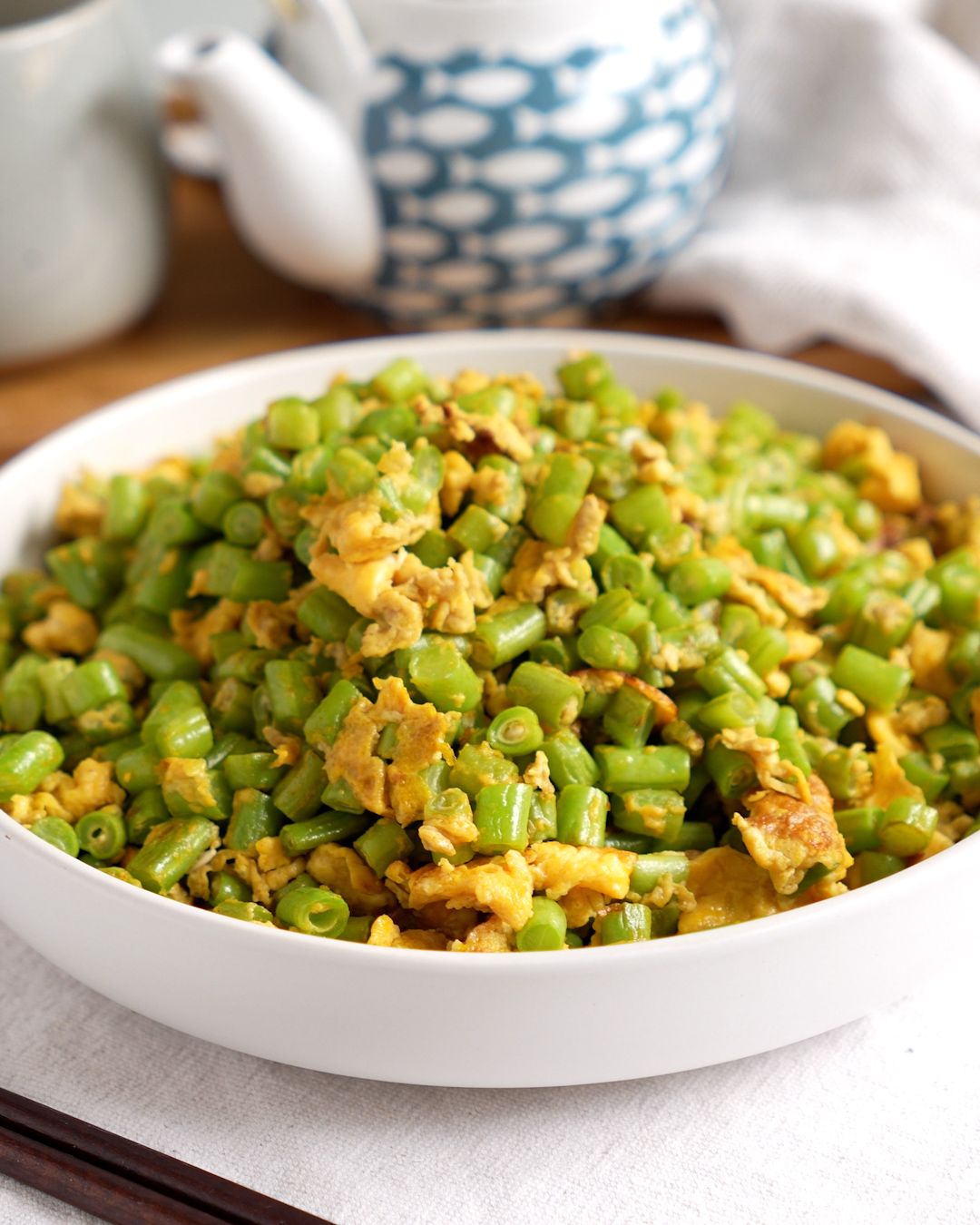 French beans with egg stir fry in a white bowl with a teapot in the background