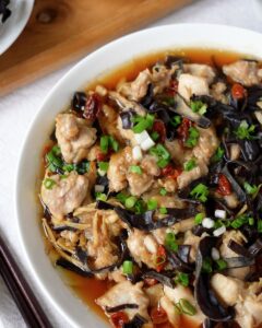 Overhead close-up of steamed chicken with black fungus, goji berries, and spring onion in a white bowl
