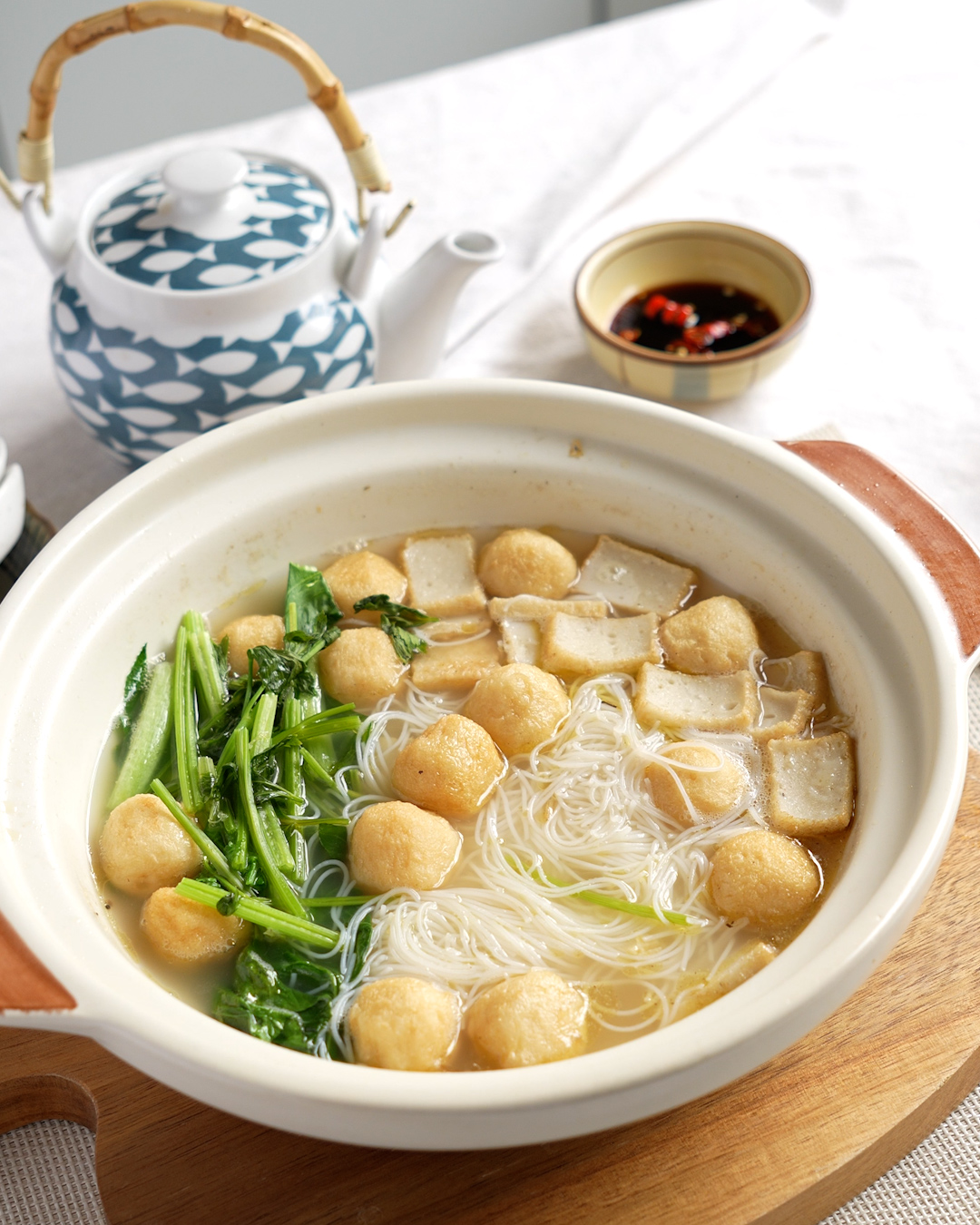 Fish ball noodle soup in a white claypot with bee hoon, fish balls, fish cake and choy sum, with chilli padi soy sauce dip and a teapot
