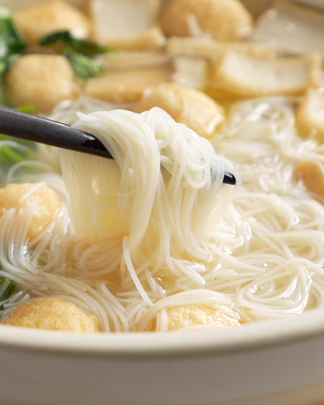 Chopsticks lifting soft bee hoon noodles from fish ball noodle soup with fish balls and fish cake in light ikan bilis broth