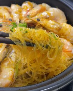 Chopsticks lifting garlic prawn glass noodles from a black pot of prawns cooked in garlic oil